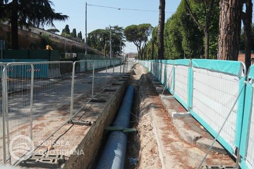 Panoramica del cantiere lungo via Tiburtina con mezzi d'opera, recinzioni e scavi aperti per la posa della nuova condotta dell'Acqua Vergine. Sono visibili tubazioni di grande diametro, macchinari e operatori al lavoro nell'area delimitata. Sullo sfondo si distinguono la carreggiata stradale e gli edifici circostanti. L'immagine documenta l'avanzamento dei lavori preparatori indispensabili alla realizzazione della futura linea tranviaria Tiburtina.